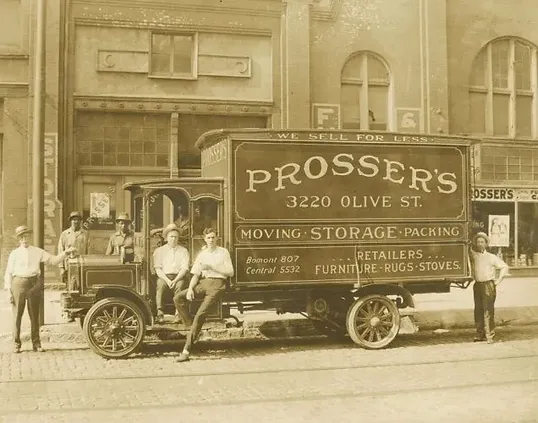 A vintage delivery truck with "Prosser's" branding is parked on a bustling city street, capturing the attention of several men standing around it. Known for stellar local moving company services in St. Louis, Prosser’s consistently earns glowing reviews from satisfied customers.